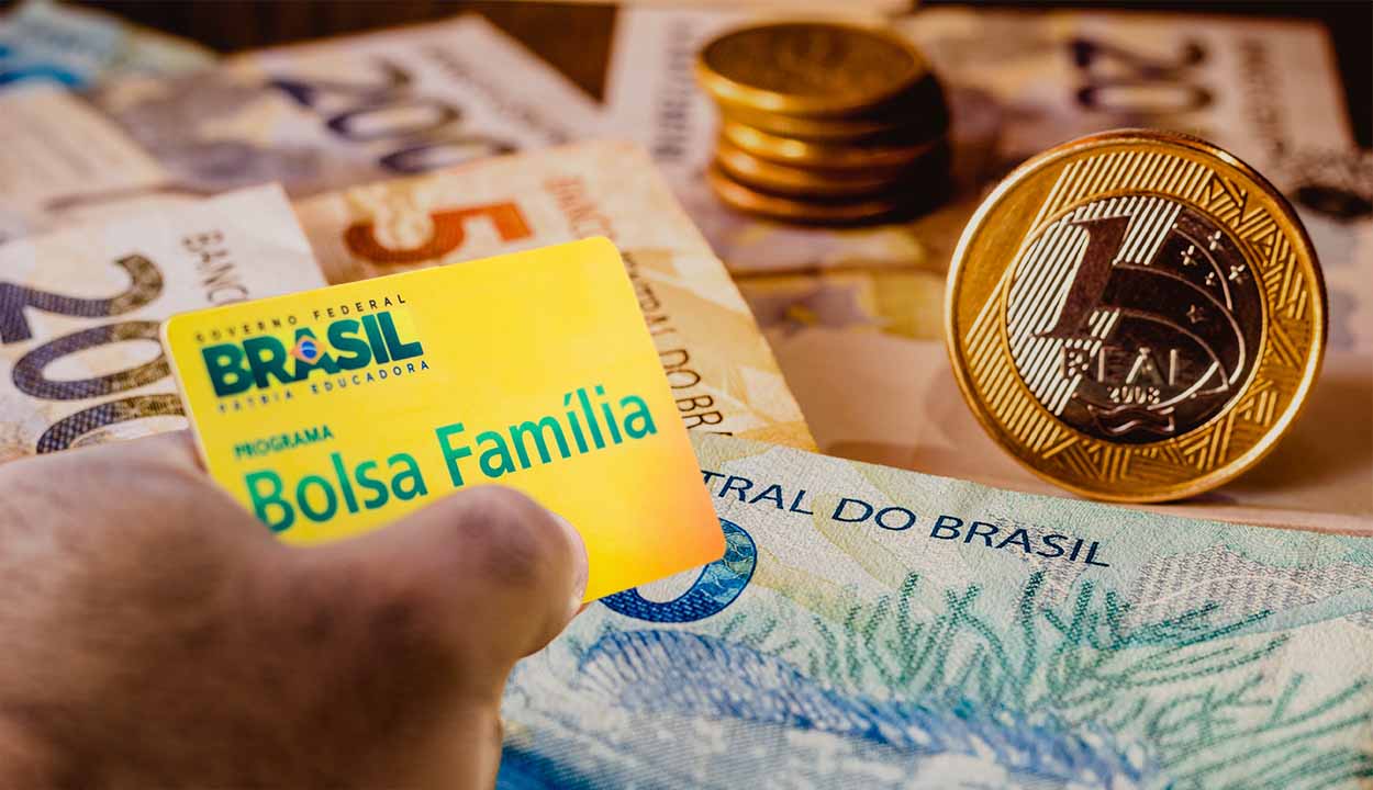MĂŁo segurando um cartĂŁo do Bolsa FamĂlia com cĂ©dulas e moedas de real ao fundo