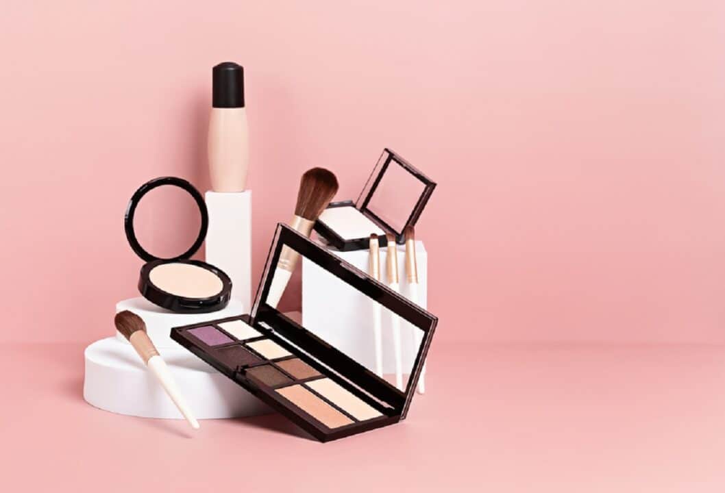 Imagem de vários tipos de maquiagem, como base, sombras, blush, e pincéis de vários tamanhos, sobre alguns degraus brancos. O fundo é rosa.