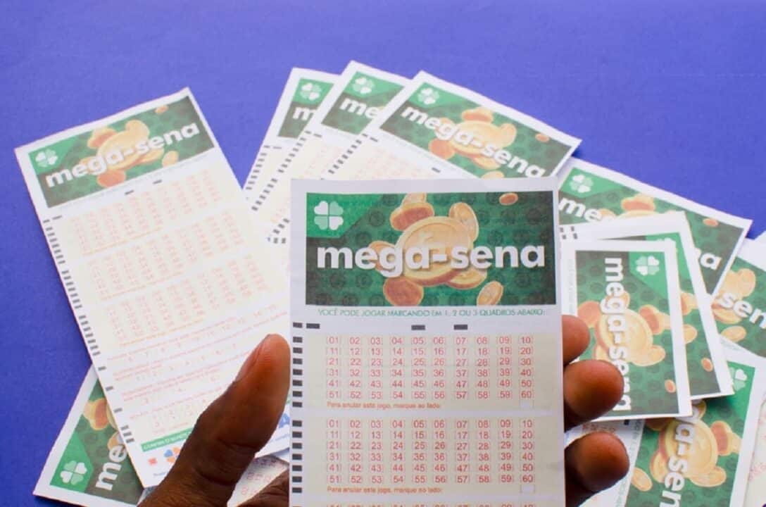 Imagem de uma mão segurando uma cartela da Mega Sena. Ao fundo, várias cartelas do concurso desfocadas.