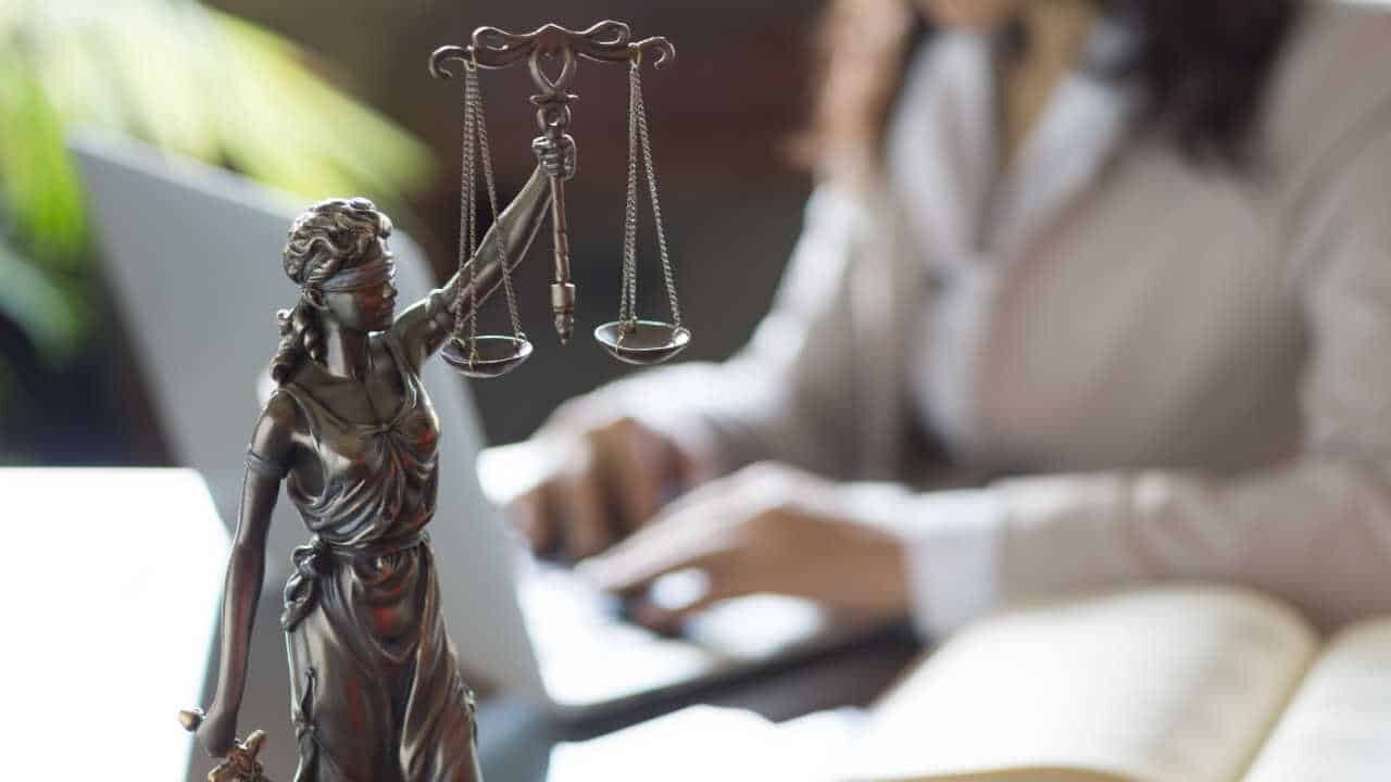 Miniatura da estátua da Justiça com mulher ao fundo enquanto digita em notebook TJ