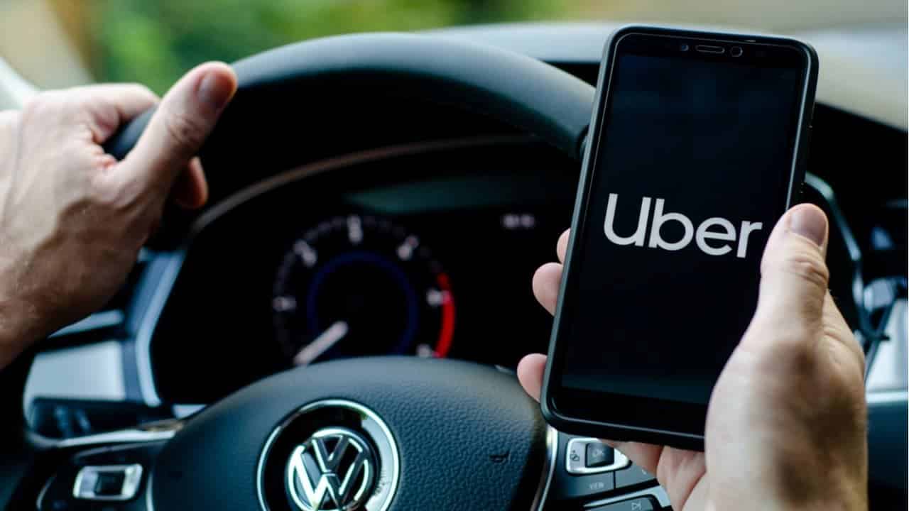 Motorista com uma mão sobre o volante e a outra segurando celular com app Uber aberto