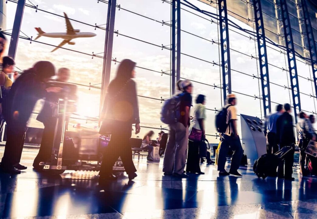 pessoas em uma fila dentro de um aeroporto. Aparece uma janela de onde se vê um avião voando.