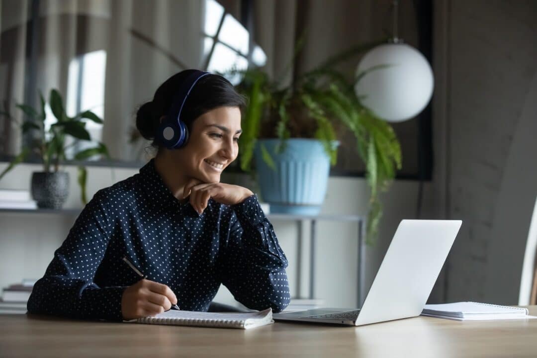 Imagem mostra uma mulher sorrindo e olhando para o computador. Ela está com fone de ouvido e segurando uma caneta para fazer anotações.