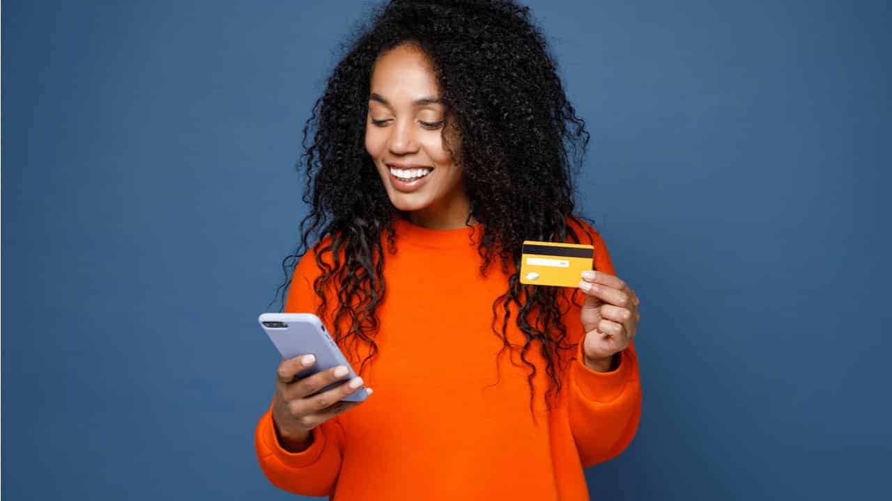 Mulher sorrindo com cartão de crédito em uma mao e celular em outra. Ela usa blusa laranja e está em um fundo azul