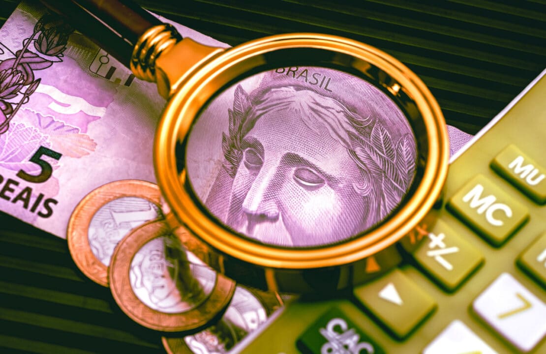Lupa sobre moedas, calculadora e nota de 5 reais