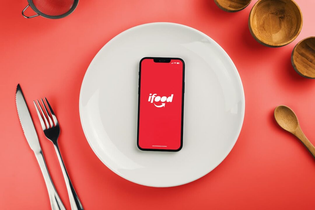 Celular com aplicativo do iFood dentro de um prato branco sobre uma mesa vermelha e talheres ao lado.