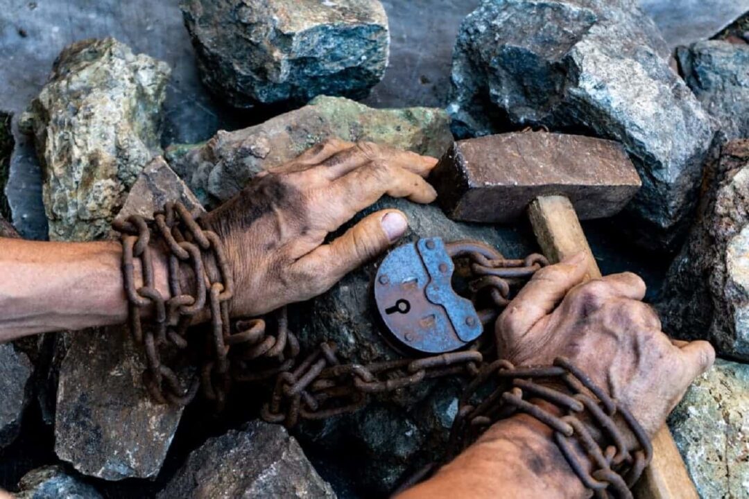 Imagem de duas mão acorrentadas sobre pedras, segurando um martelo, em analogia ao trabalho escravo