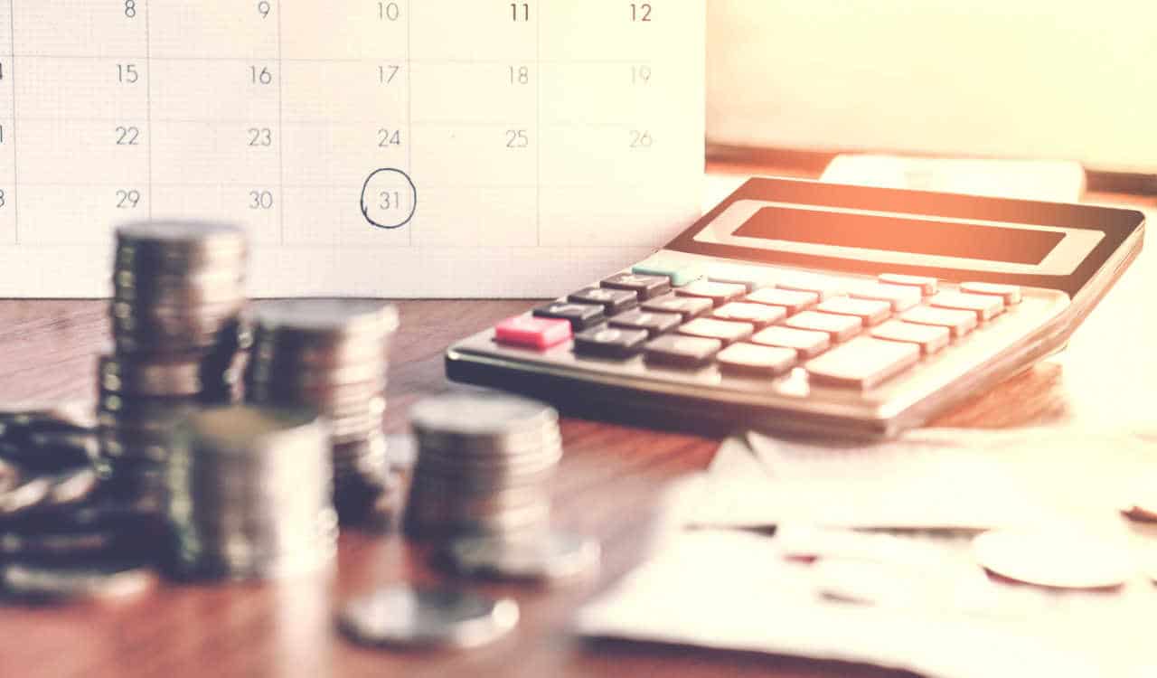 Em cima de uma mesa estão um calendário com o dia 31 marcado, uma pilha de moedas, uma calculadora e várias contas