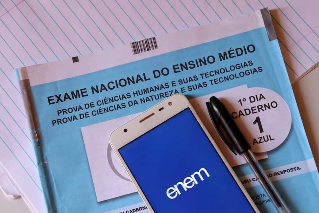 Um celular, aberto na página do Enem, e uma caneta em cima de prova do Enem (Exame Nacional do Ensino Médio)