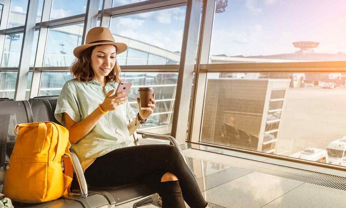 mulher no aeroporto conferindo o celular e com um copo de café na mão