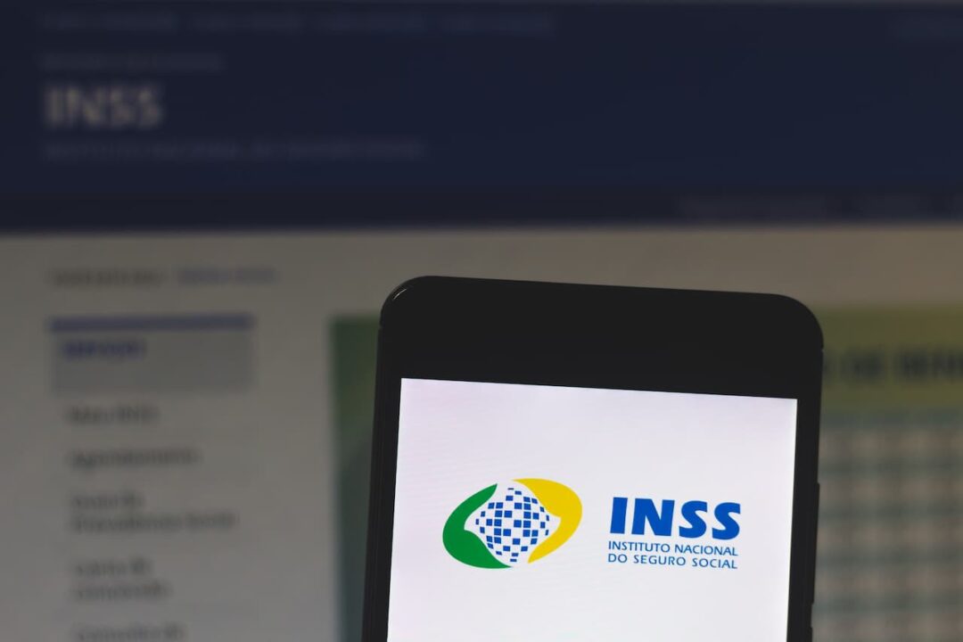 Imagem mostra, em primeiro plano, um celular com a logo do INSS. Em segundo plano, há uma tela exibindo a página do instituto