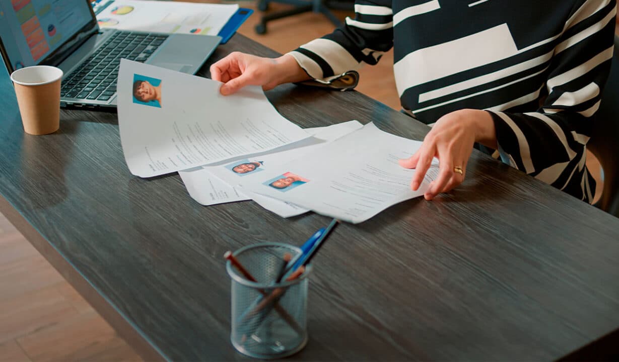 Pessoa folheando diversos currículos pousados sobre uma mesa