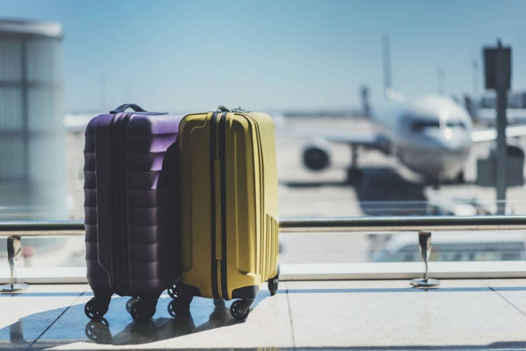 Imagem de duas malas juntas em um aeroporto. Ao fundo, um avião parado na pista.