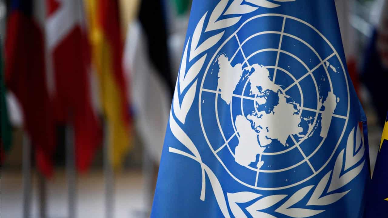 A imagem mostra a bandeira da ONU, aparentemente em um lugar fechado, em um mastro, com as bandeiras de outros países ao redor.