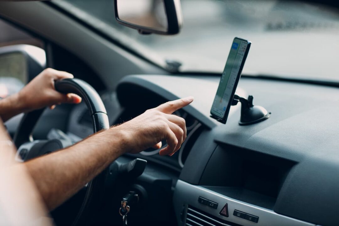 Motorista de aplicativo de transporte apontando dedo para celular enquanto segura o volante com a outra mão.