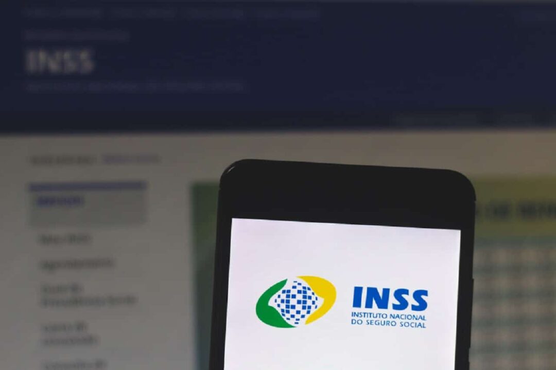 Celular com tela ligada no aplicativo do INSS, ao fundo, tela de um computador