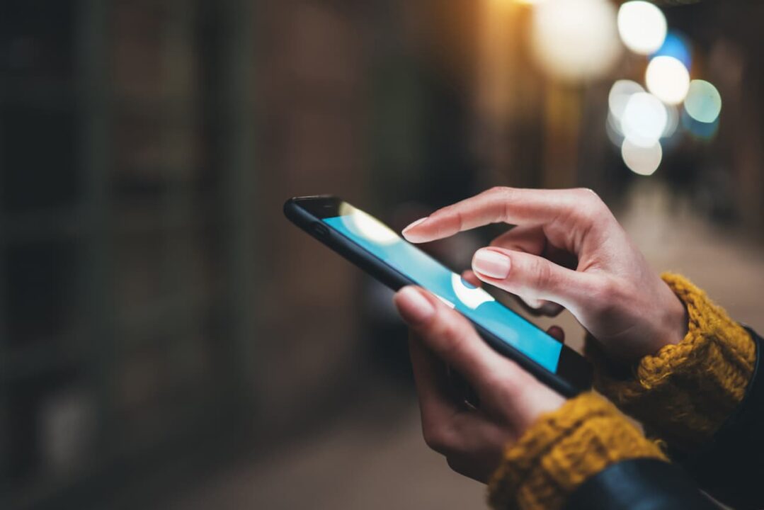 Imagem de pessoa mexendo no celular, uma mão segurando e a outra com o dedo indicador na tela. Só aparecem as mãos