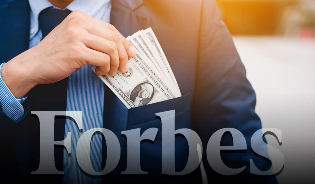 Na imagem um homem está inserindo notas de dólares no bolso externo do paletó e abaixo há a palavra "Forbes".