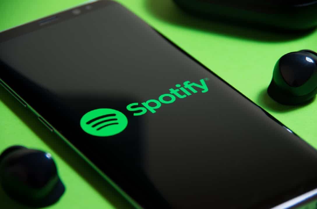 Um celular exibindo o logotipo do app Spotify em cima de uma mesa verde. Em cada lado do aparelho, tem um fone de ouvido.
