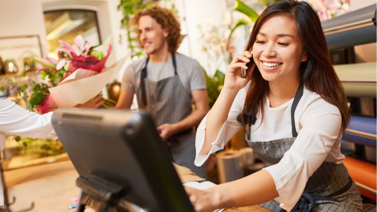 imagem de dois atendentes trabalhando em caixa de loja física, uma mulher atende o telefone enquanto usa computador, ao lado dela um homem entrega um buquê de flores para cliente