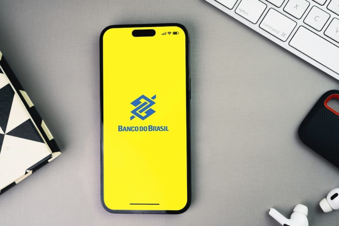 Posicionado no meio de um par de fones de ouvido e um teclado, um celular exibe a tela inicial do app do Banco do Brasil