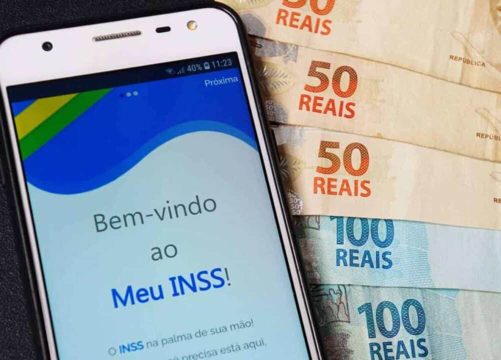 Celular acessando o aplicativo do INSS, ao lado, cédulas de cem e cinquenta reais
