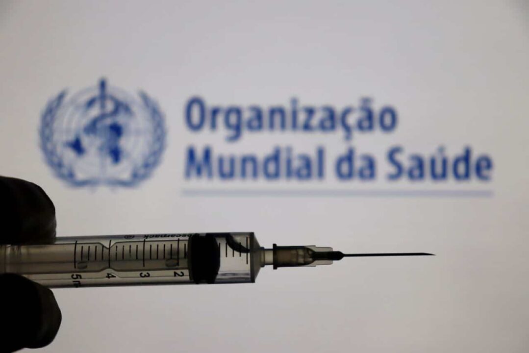 ao fundo, painel com a identidade visual da Organização Mundial de Saúde (OMS), e na frente uma vacina