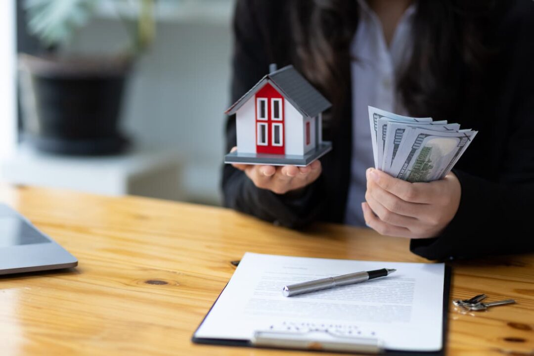 Imagem de uma pessoa segurando uma casa em miniatura e notas de dinheiro representando o aluguel de um imóvel.