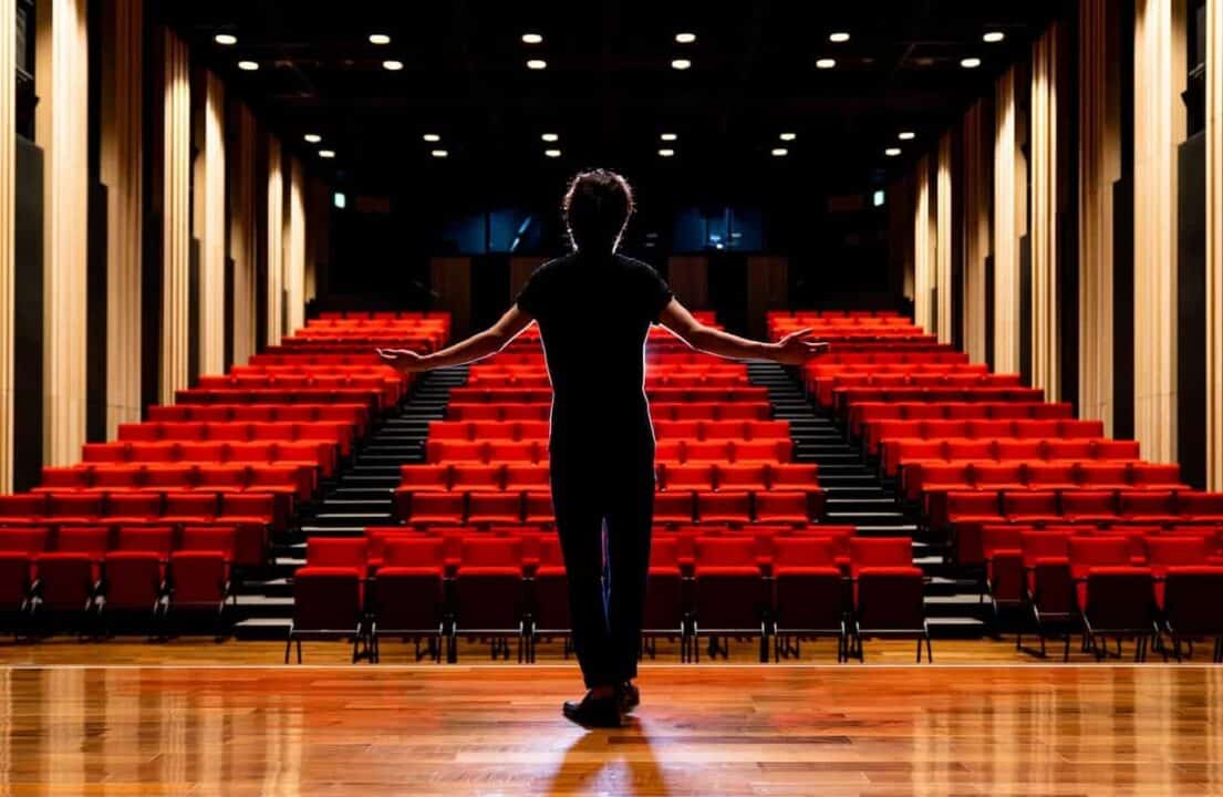 ator de braços abertos enquanto contracena em cima de palco de teatro