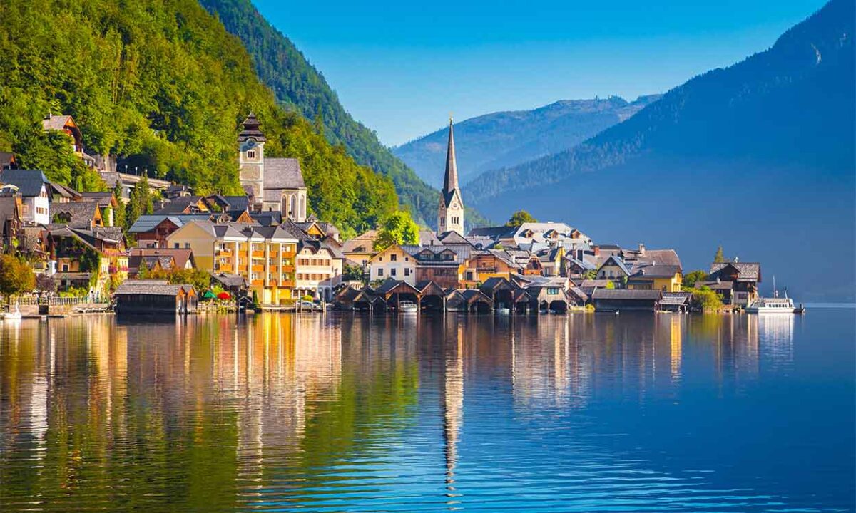 Uma cidade com aspecto antigo na beira de um lago. A cidade se reflete na agua e há montanhas ao fundo.