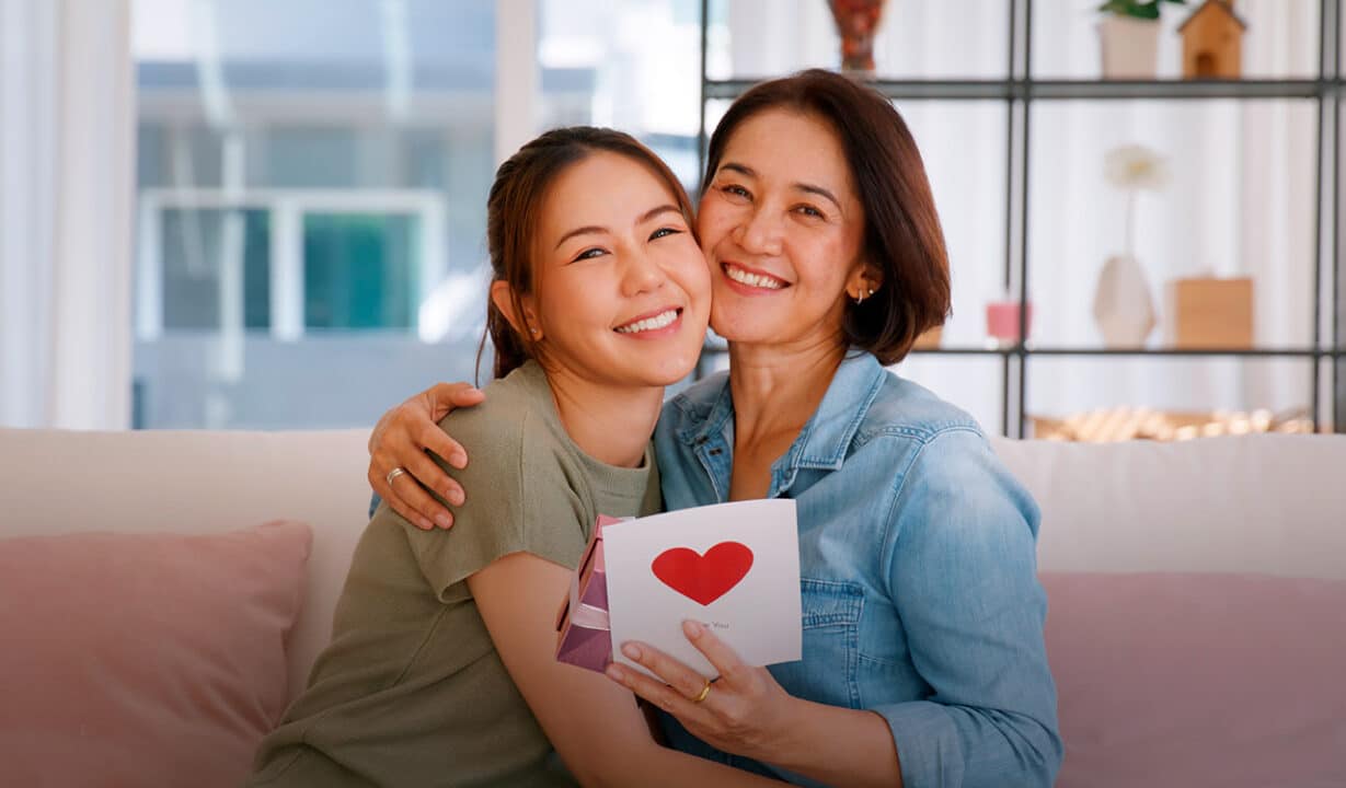 mãe e filha se abraçam, a mãe está segurando uma caixa de presentes e um cartão com um coração desenhado, escrito "I love you"