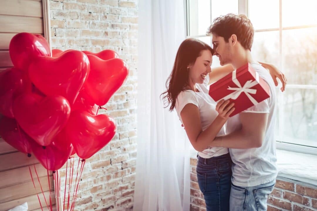 Balões de coração ao lado de um casal apaixonado se presenteando no Dia dos Namorados.