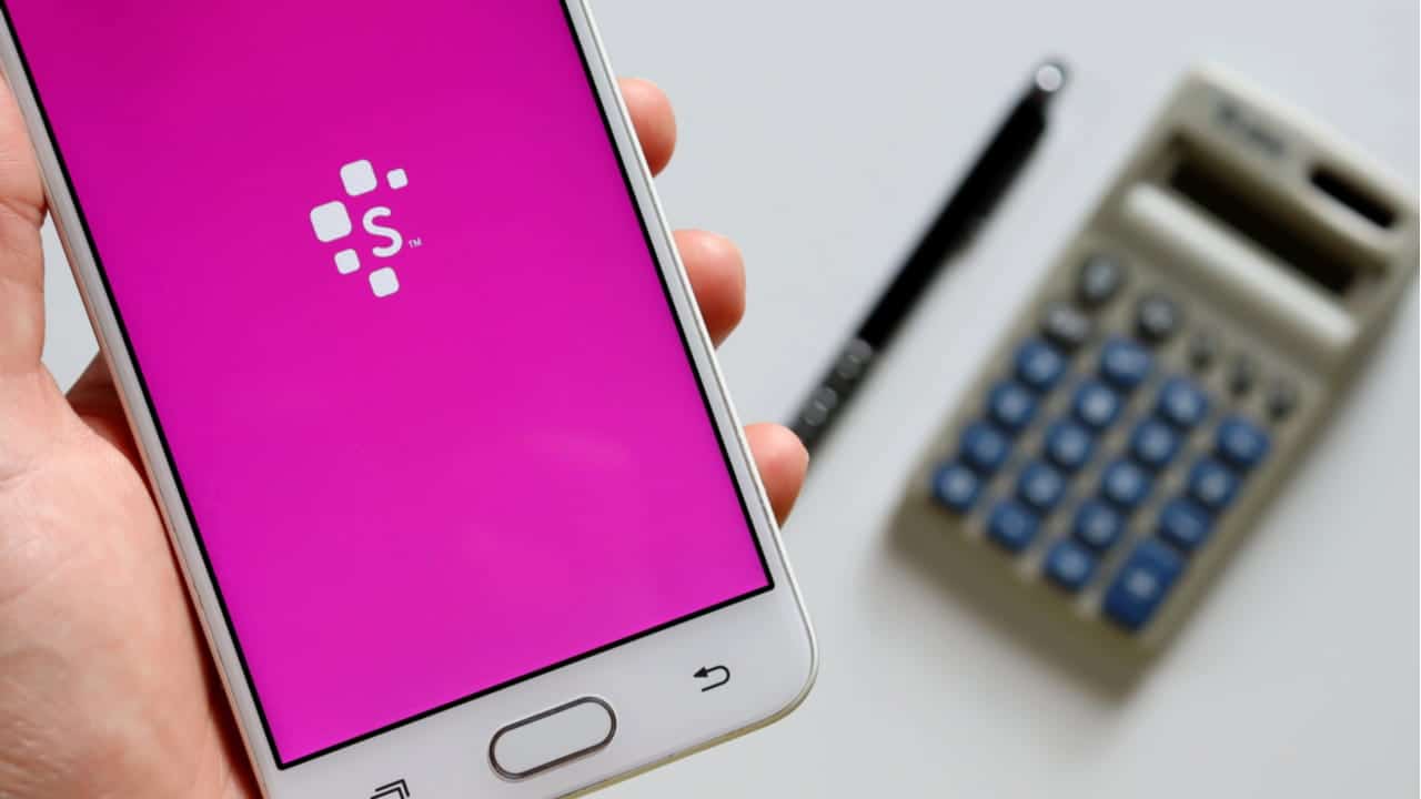 Mão segurando celular que mostra tela inicial app Serasa. Em segundo plano há uma calculadora e uma caneta posicionadas em uma superfície branca