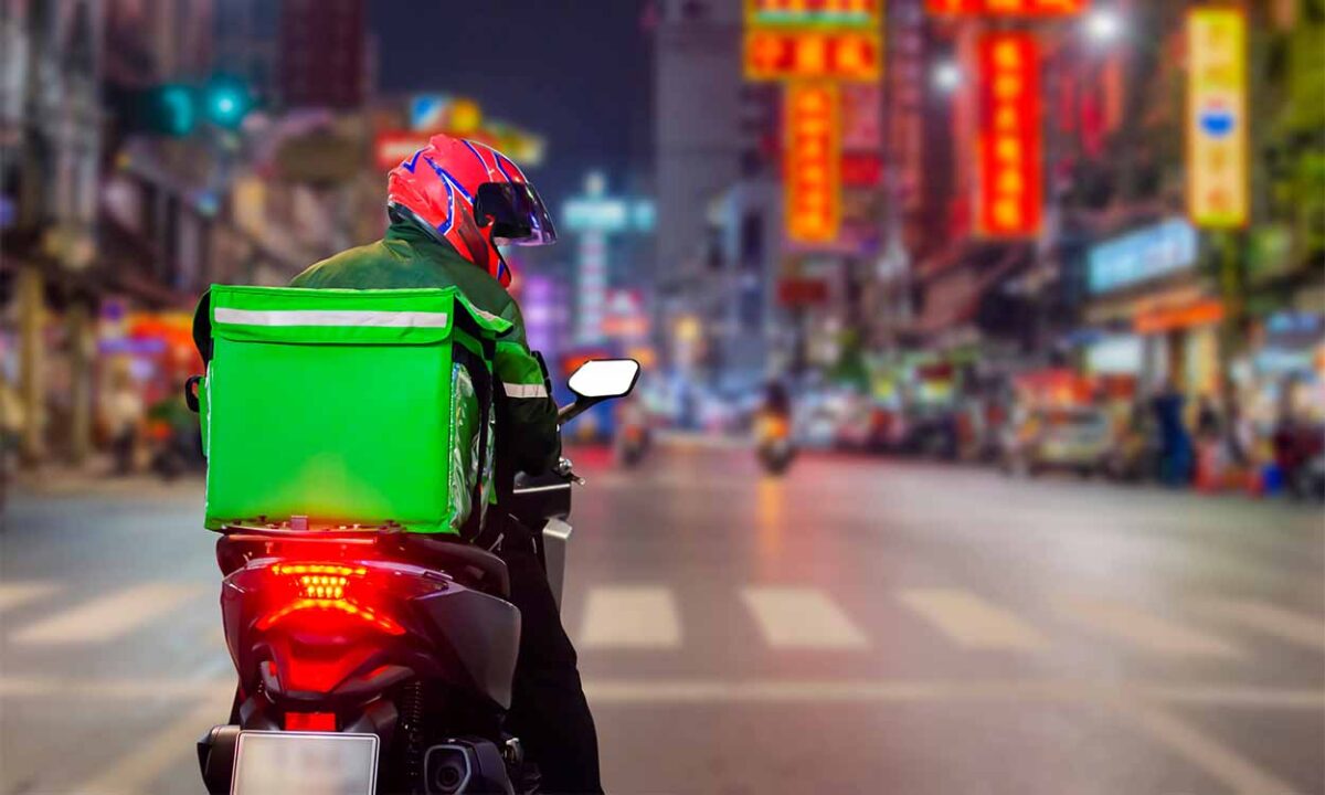 entregador de delivery dirigindo moto numa avenida, ele usa um capacete vermelho e uma bolsa verde na garupa