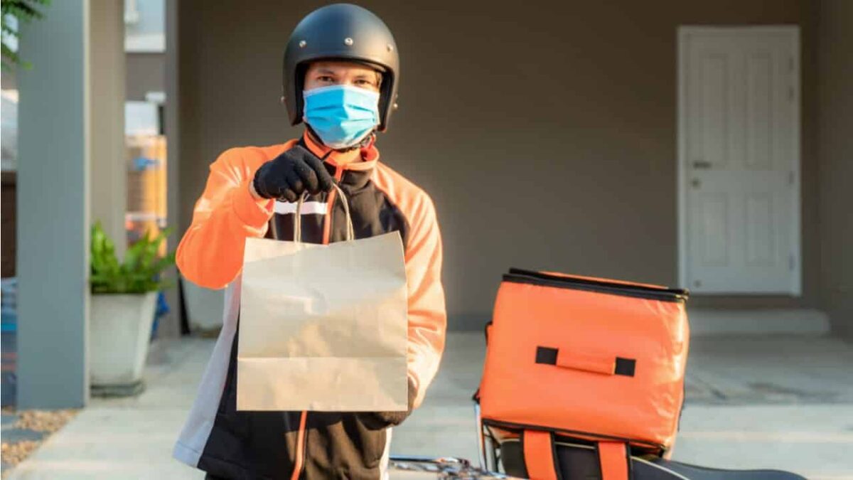Entregador de delivery ao lado de sua moto, segurando uma sacola com algum produto.