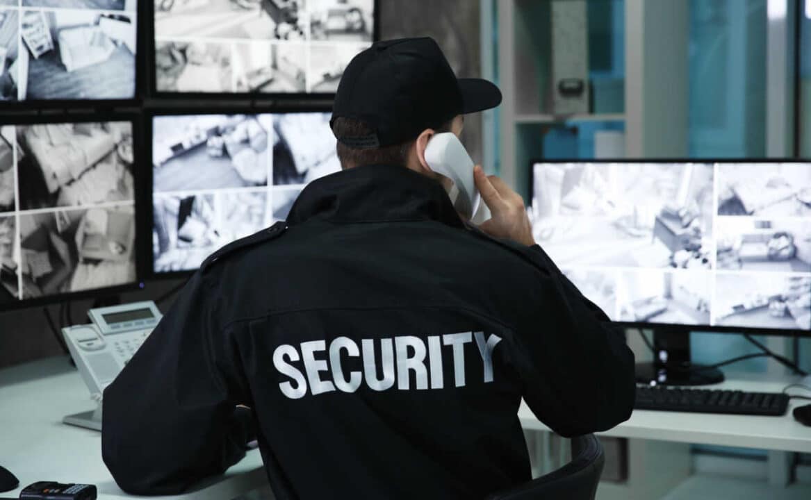 homem de costas, com um casaco escrito "segurança", fala ao telefone em uma sala cheia de telas de câmeras de vigilância