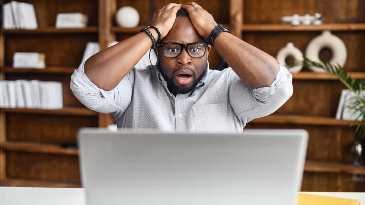 rapaz negro usando óculos coloca a mão na cabeça, com expressão de choque, enquanto olha para noticia no notebook