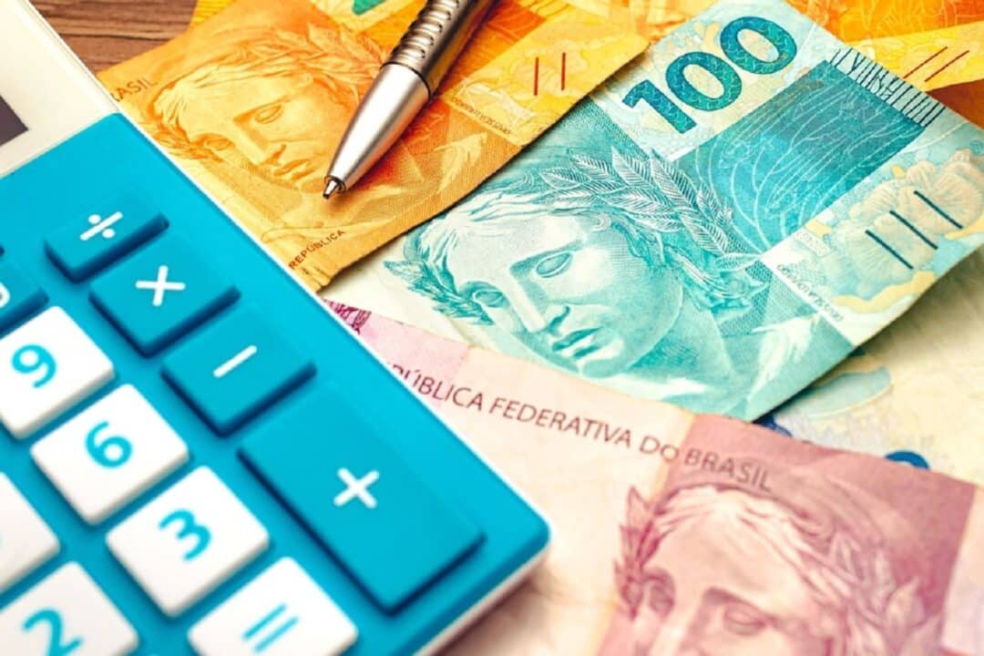 Imagem de notas de real embaixo de uma calculadora e uma caneta impostos carga tributária