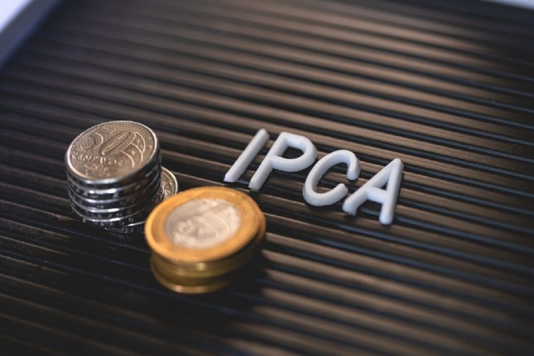 Letras brancas formando a sigla IPCA ao lado de moedas de R$ e R$ 0,50. Os objetos estão sobre uma superfície preta.