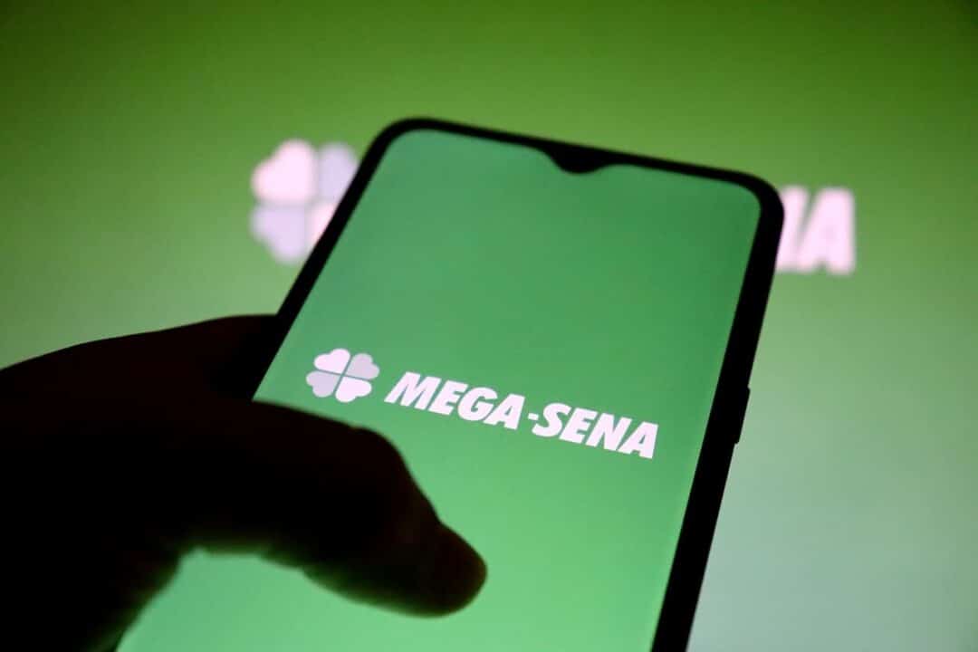 Na imagem, mão segurando celular com logo da Mega-Sena