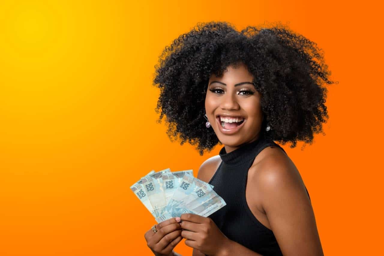 Mulher negra com expressão de felicidade segurando notas de dinheiro de cem reais.
