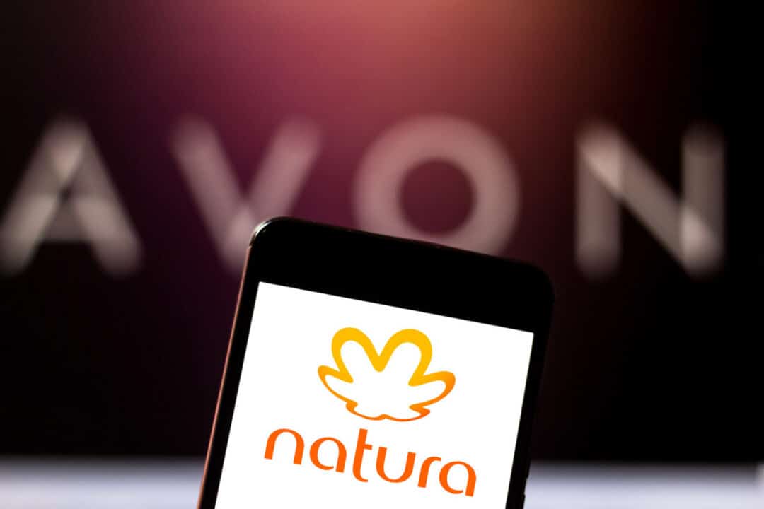 Celular com logo da Natura na tela, e de fundo, o nome da Avon
