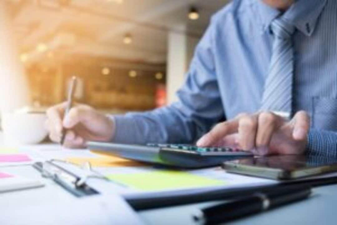 Homens calculando finanças com calculadora e caneta. Na mesa estão alguns papéis. Ao fundo, sala de escritório.