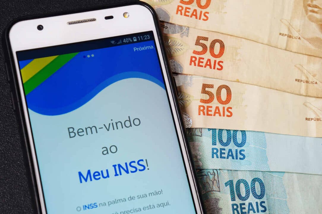 Celular acessando o aplicativo do INSS, ao lado, cédulas de cem e cinquenta reais