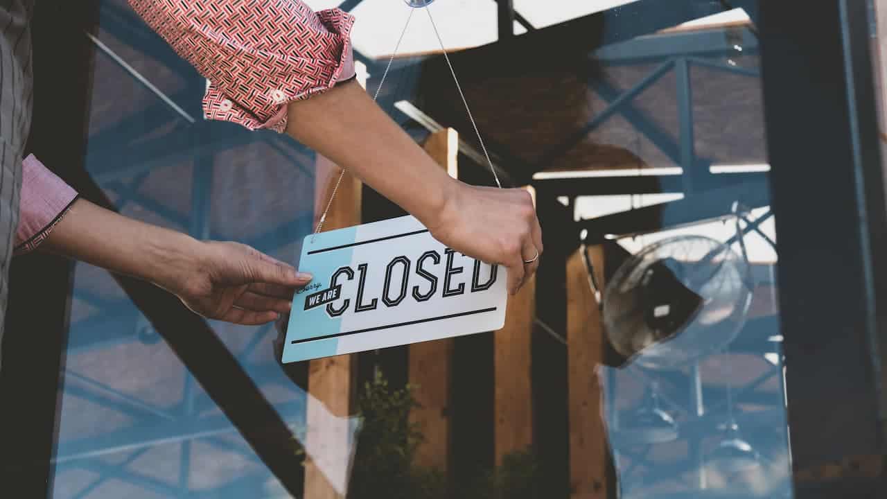 Pessoa colocando uma placa escrito "closed" na porta de um estabelecimento