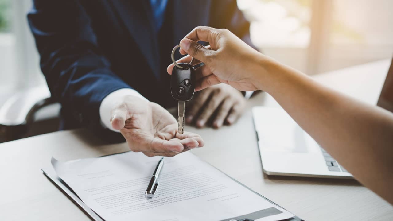 Mão esticada entregando a chave de um carro para outra mão. Abaixo, um papel que parece um documento, uma caneta e uma parte de um notebook são visíveis transferência