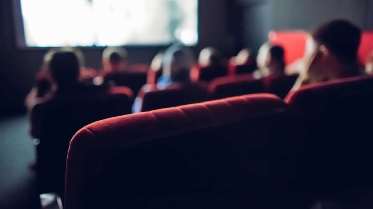 Pessoas em uma sala de cinema, com poltronas vermelhas, assistindo a um filme. A imagem é um pouco desfocada.