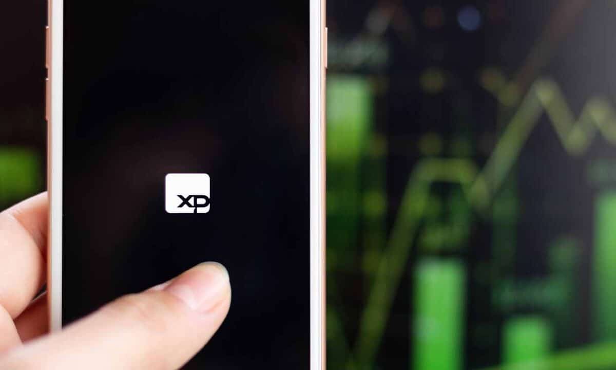 Imagem de uma mão segurando um celular com a tela na logo da XP. Ao fundo, gráficos