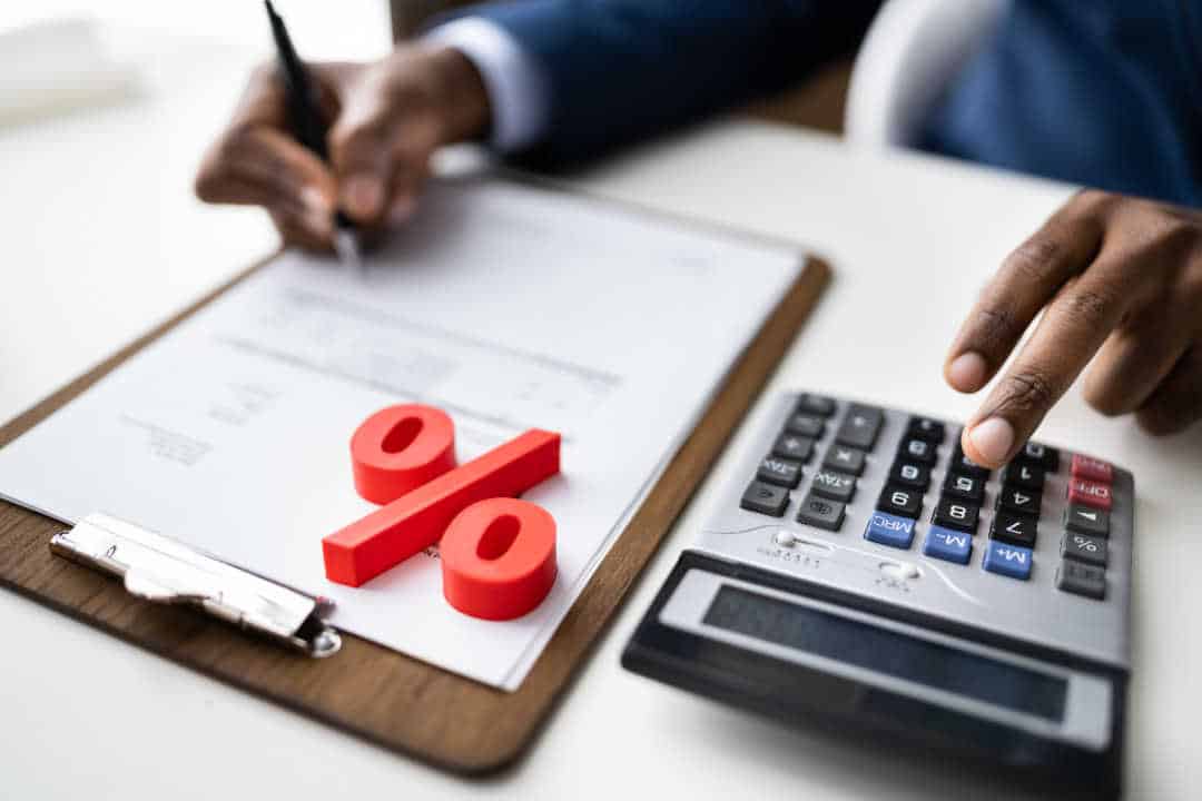 Pessoa fazendo anotações em uma prancheta e usando uma calculadora com um símbolo de porcentagem vermelho em cima dos papéis