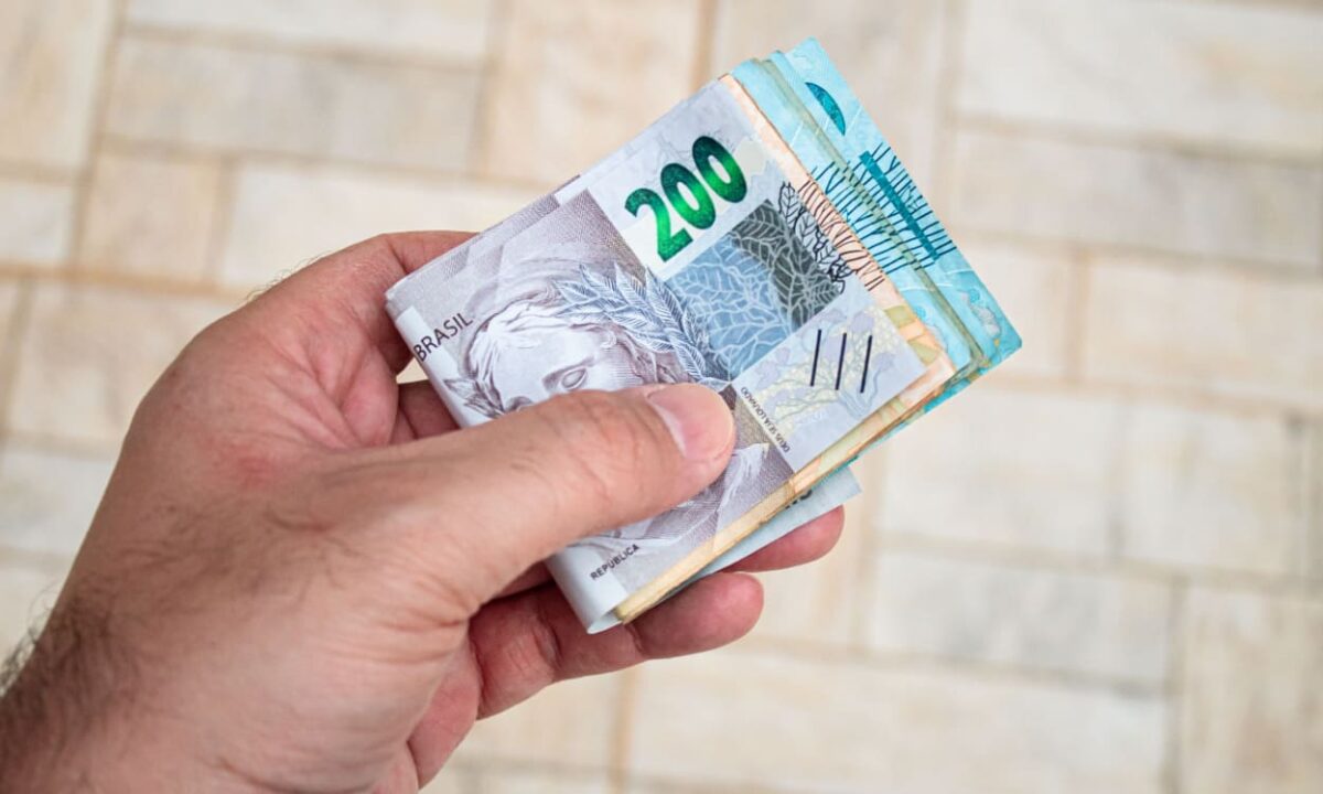 Pessoa segurando um maço de dinheiro com várias notas de Real do 13° salário, representando a antecipação do pagamento do governo.